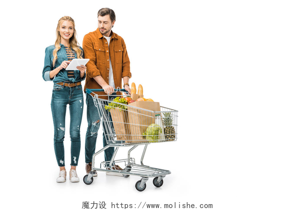 在白色背景墙前一对情侣超市在购物年轻的妇女检查购物清单上的数字平板电脑, 而她的男朋友站在附近与推车满纸袋与产品查出白色